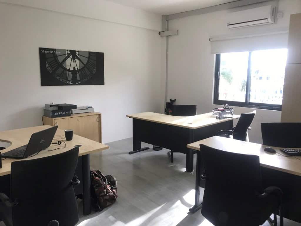 Espace de travail avec bureaux, ordinateurs et grande fenêtre lumineuse.